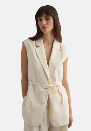 Jonge vrouw met stijlbruin haar die een mouwloos crèmekleurig blazer draagt met een dunne riem in de taille gestrikt, handen in de zakken, neutrale achtergrond.