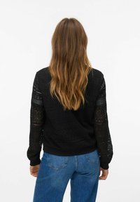 Zwarte gebreide trui met ingewikkelde patronen, lange mouwen en ribgebreide manchetten. Gecombineerd met blauwe high-waisted jeans in een ontspannen pasvorm.