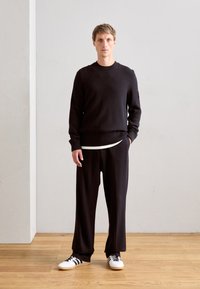 Man standing indoors wearing a black ribbed sweater, loose black trousers, white sneakers with black stripes, and a white undershirt visible.