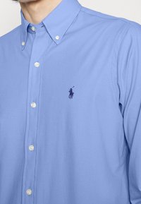 Jasnoniebieska koszula na długi rękaw z kołnierzykiem w szpic i białymi guzikami. Posiada małe fioletowe wyszywane logo na piersi.