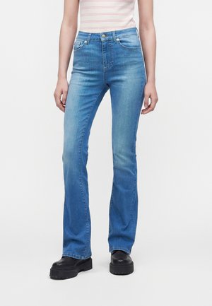 Kobieta ubrana w jasnoniebieskie jeansy bootcut, czarne buty na platformie i bladoróżowy, paskowany top, stojąca na białym tle.