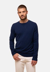 Marineblauer Rundhalsausschnitt-Pullover aus einem weichen, strukturierten Stoff mit gerippten Bündchen und Saum, kombiniert mit hellen Hosen.