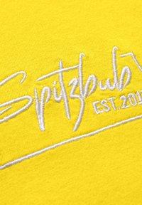 Gelbes Gewebe mit einer weichen Textur zeigt weißen gestickten Text, der "Spitzbub EST. 2018" in einer lässigen Schriftart liest.