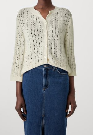 Person trägt eine cremefarbene, leichte Strickjacke mit Knöpfen und einen hoch taillierten blauen Jeansrock mit Frontschlitz.