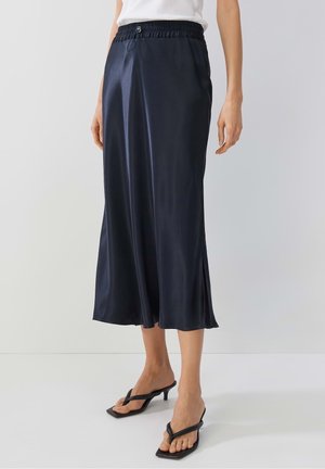 Kobieta nosząca granatową satynową spódnicę midi z elastycznym pasem i bocznym rozcięciem, w połączeniu z czarnymi sandałami na obcasie i białą bluzką.