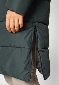 Tamnozeleni zimski kaput od puf materijala s kvadratnim uzorkom, s bočnom zip džepom. Materijal izgleda lagano i otporno na vodu.