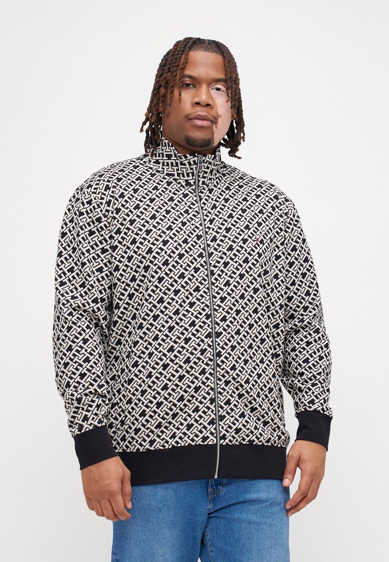 Veste zippée avec un motif géométrique noir et blanc, fabriquée dans un tissu doux, avec un col montant et des poignets noirs contrastants.