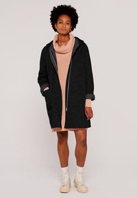 Svart oversized jacka med grå kant, med stora fickor och en framdragkedja. Bärs över en rosa stickad tröja och krämfärgade högskor.