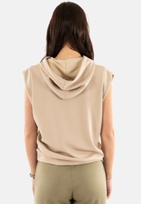 Top sin mangas con capucha en color beige, de tela suave, con un corte relajado y detalles de costura cerca de los hombros. Combinado con pantalones en color verde oliva.