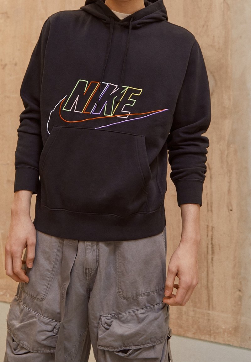 Black hoodie with multicolored Nike logo, features kangaroo pocket and ribbed cuffs. Worn with light gray cargo pants.