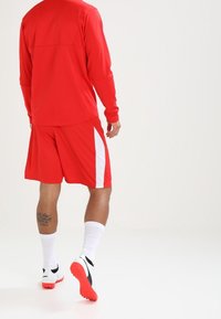 Mann in roter Sportbekleidung mit weißen und schwarzen Schuhen, der auf einer weißen Fläche mit weißem Hintergrund steht.