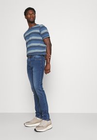 T-shirt a righe blu, jeans in denim slim-fit e sneakers beige. La maglietta presenta diverse tonalità di righe blu e un collo rotondo.