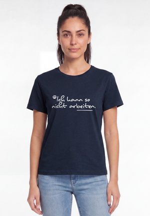 Frau mit dunklen Haaren im Pferdeschwanz trägt ein marineblaues T-Shirt mit weißem deutschen Text "Ich kann so nicht arbeiten" und hellblaue Jeans.