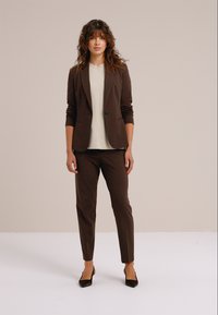 Sacou maro croită pe comandă peste un top crem, asortat cu pantaloni maro conici și pantofi negri cu vârf ascuțit. Material neted cu design structurat.