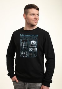Mann in schwarzem Sweatshirt, das sechs Szenen aus der Serie "Wednesday" zeigt, darunter ein Mädchen mit Zöpfen und gothischen visuellen Elementen.
