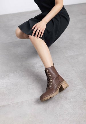 Botas de tobillo de cuero marrón con ojales dorados, un tacón grueso y una suela robusta; colocadas sobre un fondo gris neutro.