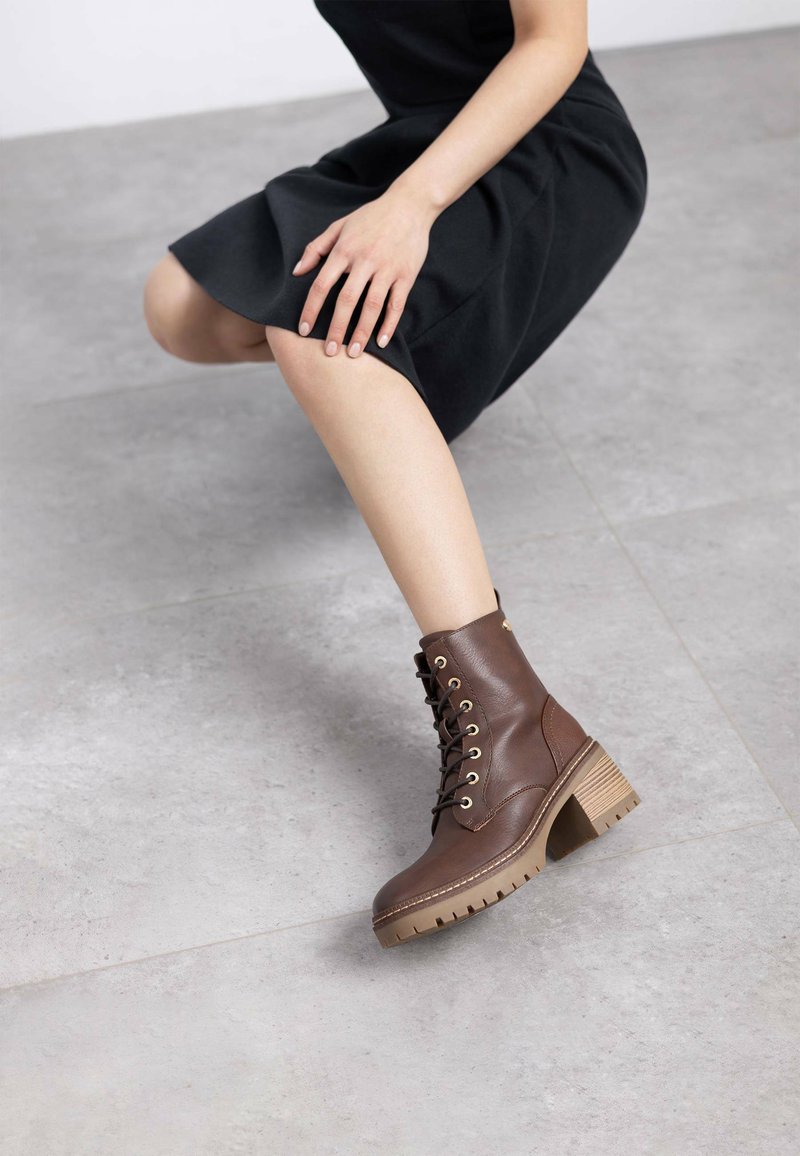 Botas de tobillo de cuero marrón con ojales dorados, un tacón grueso y una suela robusta; colocadas sobre un fondo gris neutro.