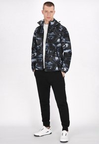 Sort og blå mønstret jakke med høj krave, båret over en hvid skjorte, parret med sorte bukser og hvide sneakers.