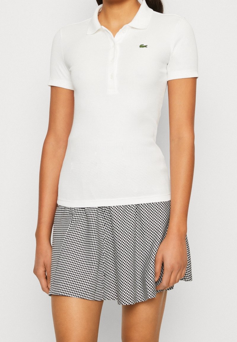 Femme portant un polo blanc cintré à manches courtes et une jupe plissée pied-de-poule noire et blanche, debout devant un fond uni.