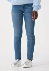 Chude niebieskie dżinsy o subtelnej teksturze, w stylu średniego wzrostu, zwężane nogawki, zestawione z metalicznymi srebrnymi trampkami.