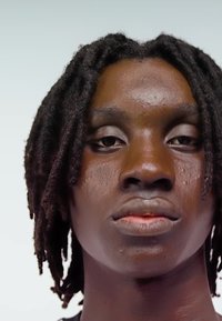Záběr zblízka na osobu s tmavou pletí a krátkými dredy na světle šedém pozadí, která se dívá přímo před sebe s neutrálním výrazem.