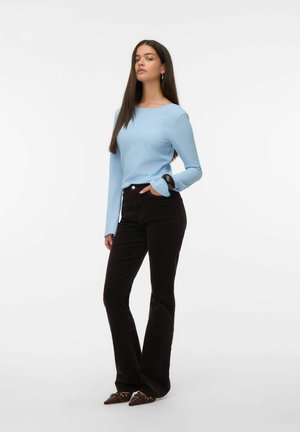 Vrouw met lang, donker haar die een lichtblauw, textuurgegeven, langemouwen shirt, zwarte, hoge taille broeken en puntige, bruine schoenen met studs draagt.