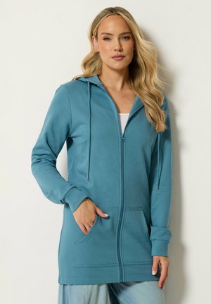 Kvinde med langt blond hår iført en turkis lynlåstrøje med forlommer og snører over lyseblå jeans mod en ensartet lys baggrund.