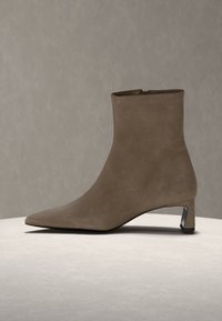 Μποτάκι αστραγάλου από καφέ σουέντ με μυτερή μύτη, κοντό τακούνι τύπου block και φερμουάρ στο πλάι, σε ουδέτερο φόντο και επιφάνεια.