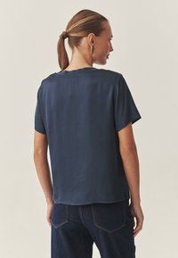 Marineblaues kurzärmliges Shirt aus glattem Stoff, mit rundem Ausschnitt und seitlichen Schlitzen, kombiniert mit dunkelblauer Jeans.