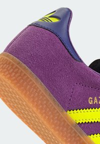 Fioletowy zamszowy obcas trampka z żółtymi zygzakowatymi paskami, przezroczystą gumową podeszwą oraz granatową skórzaną końcówką obcasa z wytłoczonym logo.