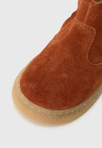 Stivale in suede marrone con suola in crepe beige, cuciture visibili e design della punta arrotondata su uno sfondo bianco.