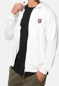 Hvid hættetrøje med lynlås, forlomme, snoretræk og et logo i læder, kombineret med en sort skjorte under.