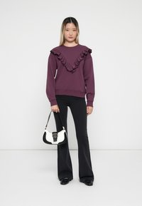 Sweatshirt violet avec encolure volantée, pantalon noir évasé et sac à main blanc avec accents noirs. Texture lisse et design décontracté.