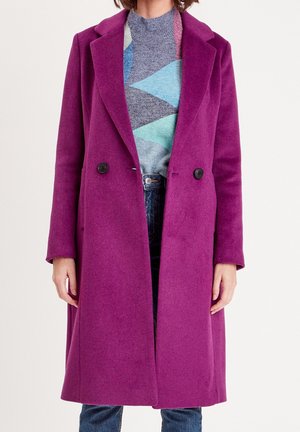 Long manteau violet à double boutonnage porté sur un pull géométrique bleu et gris, associé à un jean bleu foncé.