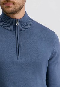 Blauer Strickpullover mit hohem geripptem Kragen und halbem Reißverschluss. Glatte Textur, taillierte Passform und minimale Nähdetails.