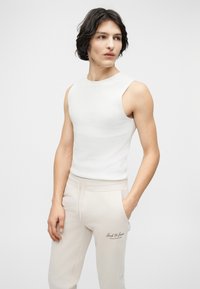 Νέος άνδρας με μαύρα μαλλιά, φορώντας λευκό αμάνικο τοπ και ανοιχτό μπεζ φόρμα με το λογότυπο "Jack & Jones", στέκεται με το ένα χέρι στην τσέπη.