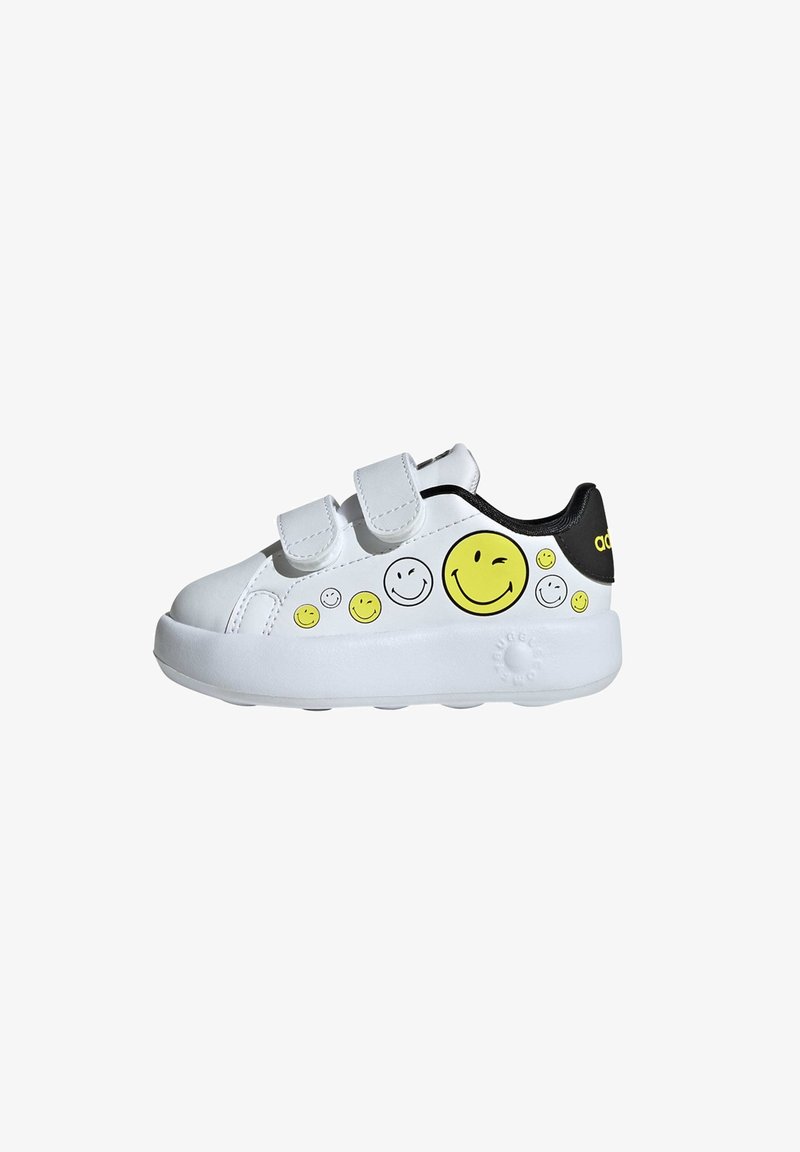 White synthetic sneaker with two Velcro straps, black accents, and yellow smiley face designs on the side. Thick white sole with textured grip.