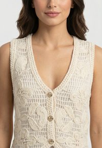 Mujer con un chaleco de ganchillo crema sin mangas con patrones florales y botones dorados, mostrado desde los hombros hasta la mitad del torso.