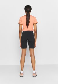 Žena s dlouhými tmavými vlasy svázanými do culíku, oblečená v broskvovém tričku, černých šortkách, bílých ponožkách a růžových teniskách, stojící před jednoduchým pozadím.