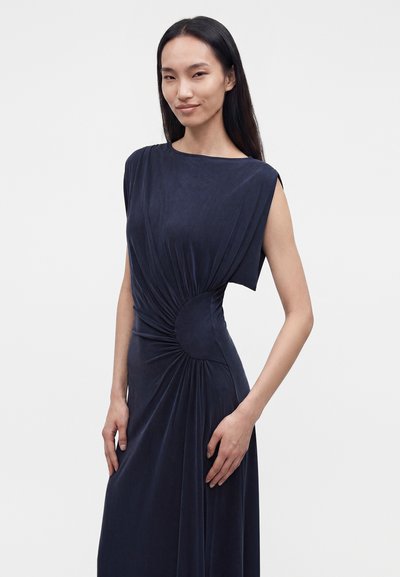 Femme aux longs cheveux foncés portant une robe bleu marine sans manches avec des fronces à la taille, debout devant un fond clair uni.