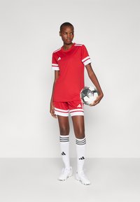 Červená športová dres s bielymi pruhmi a logom, zodpovedajúce šortky, biele ponožky s čiernymi pruhmi, drží čierno-biely futbalový loptu.
