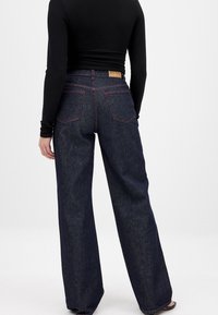 Temno denim široke hlače z rdečim šivanjem, usnjenim logotipom na hrbtni strani in dvema zadnjima žepoma. Imajo sproščen kroj.