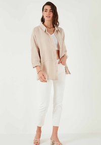 Chemise légère beige à boutons avec un tissu texturé, manches retroussées et fentes sur les côtés, assortie à un pantalon blanc court et des sandales à lanières.