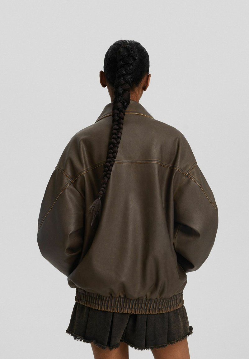 Chaqueta bomber de cuero marrón con un corte relajado, dobladillo elástico, costuras visibles y un gran cuello. Llevada sobre una falda negra con pliegues.