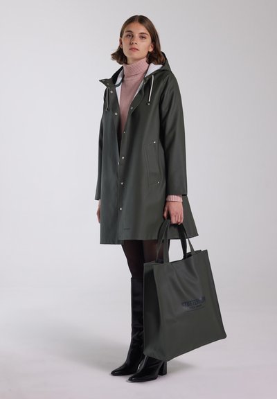 Donkergroene waterdichte regenjas met een капюшон, drukknoopsluitingen en een voorzak; gecombineerd met een bijpassende tote bag en zwarte kniehoge laarzen.