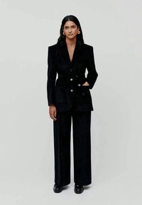 Zwarte fluwelen blazer met een ceintuur in de taille, dubbelgeknoopte knopen en scherpe revers, gecombineerd met brede zwarte broekspijpen en glanzende schoenen.