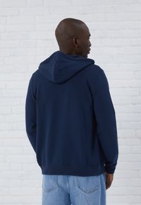 Felpa con cappuccio blu navy in tessuto morbido. Presenta una vestibilità comoda, tasca a marsupio e polsini e orlo a coste. Indossata con jeans azzurri.