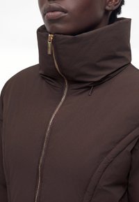 Gros plan d'une personne portant une doudoune marron foncé avec un col rembourré haut et une fermeture éclair dorée partiellement ouverte.