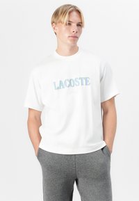 Biała bawełniana koszulka z krótkim rękawem, z kolorowym logo "LACOSTE Paris" na piersi. Noszona z szarymi spodniami dresowymi.