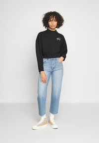 Sudadera negra recortada con mangas largas, con un logo. Combinada con jeans rectos de color azul claro y zapatillas beige de caña alta.